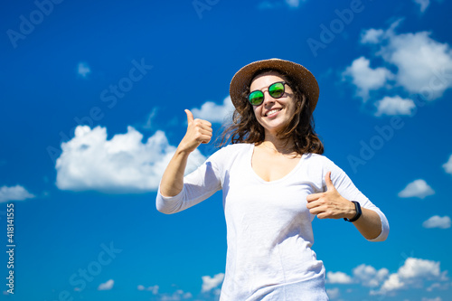Emotional smiling brunette woman showing thumb up approval like gesture, wearing summer hat, sunglasses, white clothes. Support satisfied recommend the best. Standing on blue sky background