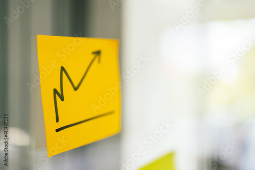 Close up of finance business graph going up that draw on yellow sticky paper on glass board with blur business people in background with copy space. The increasing business progress discussion plan. 