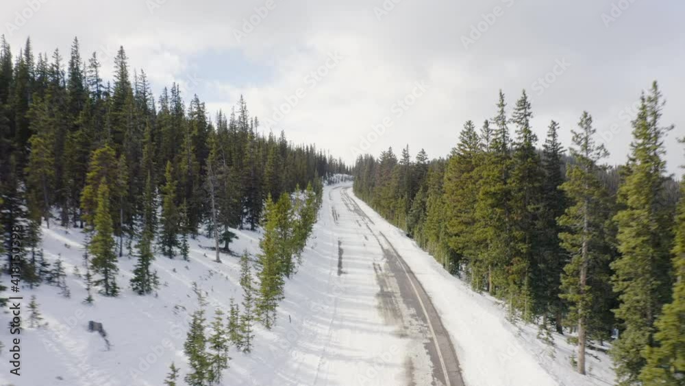 Low aerial reveal backward of sunny and snow covered back roads in the Colorado mountains surrounded by bright green pine tree forests with blue skies Low aerial reveal backward of sunny and snow covered back roads in the Colorado mountains surrounded by bright green pine tree forests with blue skies