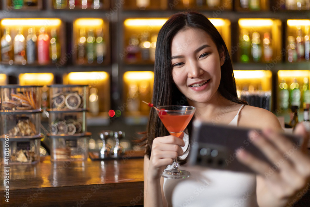Young Asian woman takes a photo of herself or Selfie with a smiling face and drinks a cocktail in a vintage bar, Relaxing activities after work or hangouts, Place of entertainment for teens.