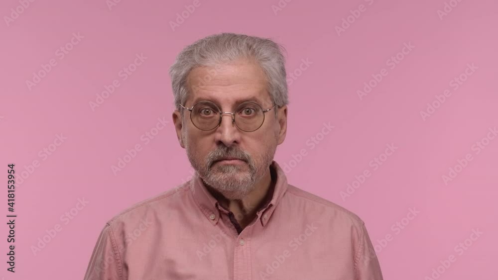 Portrait of an elderly man with glasses looks seriously at the camera, then smiles and making sign ok. Gray haired pensioner grandfather posing on pink studio background. Close up. Slow motion. Portrait of an elderly man with glasses looks seriously at the camera, then smiles and making sign ok. Gray haired pensioner grandfather posing on pink studio background. Close up. Slow motion.