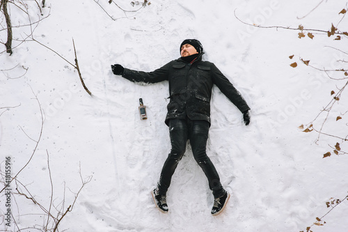 A very drunk homeless man in a black coat and hat lies, asleep on his back on white snow in a cold frosty winter with a bottle of alcohol. Photography, concept.