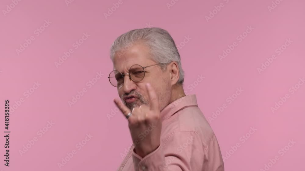 Portrait of elderly man with glasses making rock gesture, enjoying life and laughing. Gray haired pensioner grandfather with beard wearing shirt posing on pink studio background. Close up. Slow motion Portrait of elderly man with glasses making rock gesture, enjoying life and laughing. Gray haired pensioner grandfather with beard wearing shirt posing on pink studio background. Close up. Slow motion