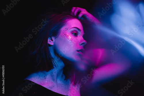 Stylish gentle portrait of a young woman in trendy makeup with glitter on a dark background posing for the camera in purple light with blue highlights, looking away.