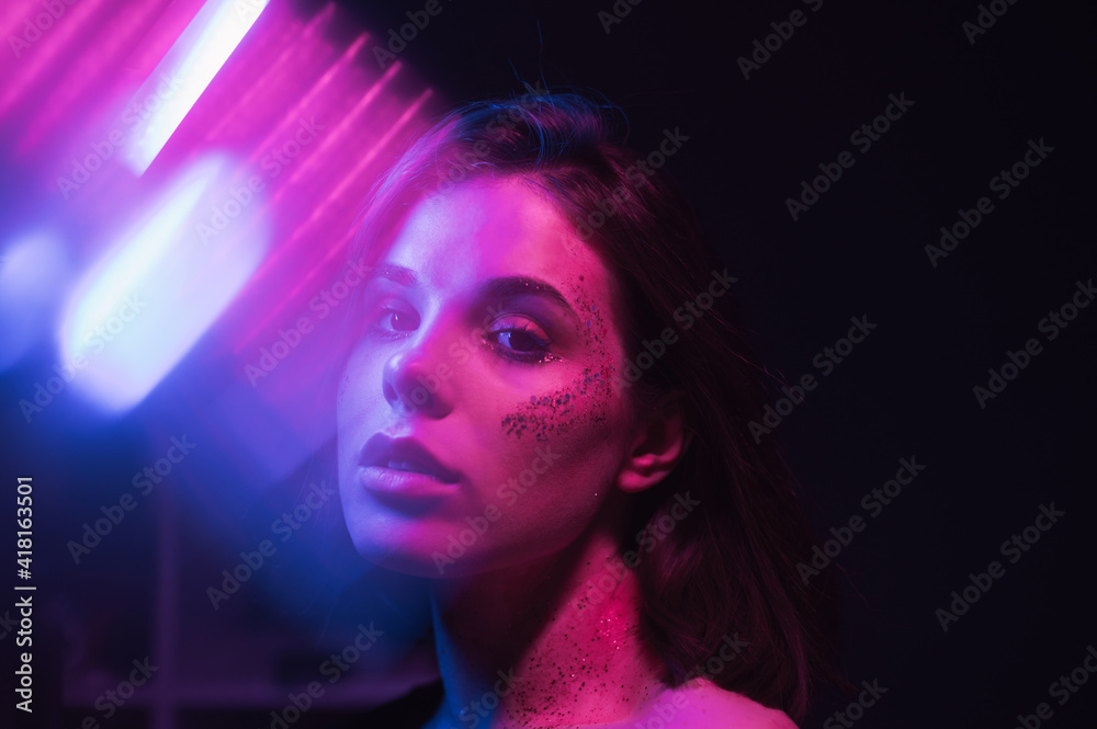 Cyberpunk photo of attractive woman in colored light on dark background with glitter on face posing at camera in blue and purple light with glare.