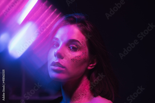 Cyberpunk photo of attractive woman in colored light on dark background with glitter on face posing at camera in blue and purple light with glare.