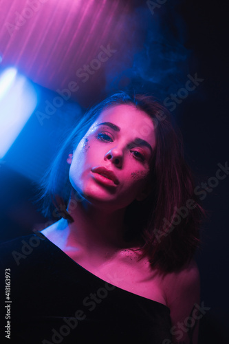 Beautiful night portrait of a woman at a party in colored light with purple and blue in the smoke, looking at the camera with a serious face. Vertical.