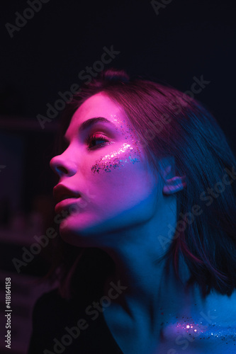 Fashion portrait of a woman with bright makeup with glitter in colored light in a dark room dancing at a party and looking away. Vertical