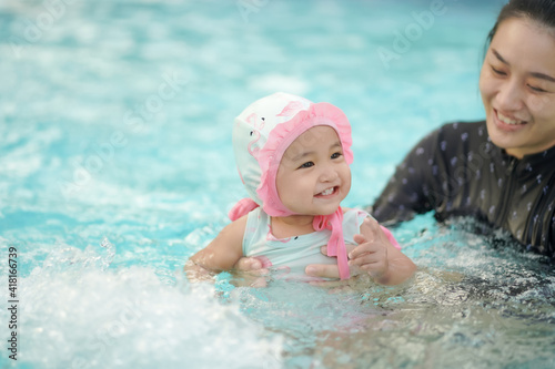 Asian cute baby girl in swimsuit holding by her young mother enjoying in the swimming pool with copy space