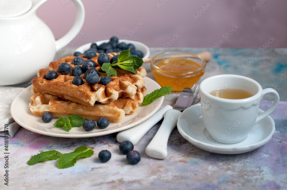 Belgian waffles with honey, blueberries, bananas, and mint on a light grey background. Breakfast with a croissant and a cup of green tea with lemon