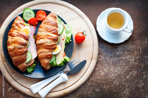 Croissant sandwich with ham, cheese, fresh salad, tomatoes, eggs and cucumber on wooden background. Breakfast with fresh croissant sandwich. Top view
