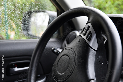 Car driver steering wheel with control panel buttons. Black dashboard and black leather covered wheel of new automobile and blurred background of raindrops on windows © Ninel