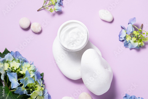 Creative composition with organic moisturizing cream on the podium made with decorative white stones and fresh hydrangea flowers on light lilac background top view. Skin care products.