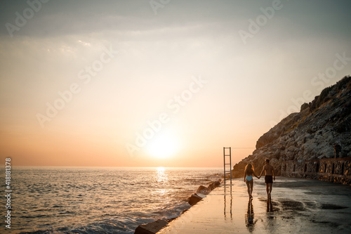 A loving couple walks along the beach by the sea. Young family at sunset by the Mediterranean Sea. Vacation concept. A woman in a swimsuit and a man in shorts at sunset by the sea. Selective focus. © Дмитрий Ткачук
