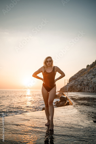 Beautiful young woman in a black swimsuit alone on the beach by the sea at sunset. An adult slender girl resting on the ocean shore at sunset. Summer vacation concept. Selective focus