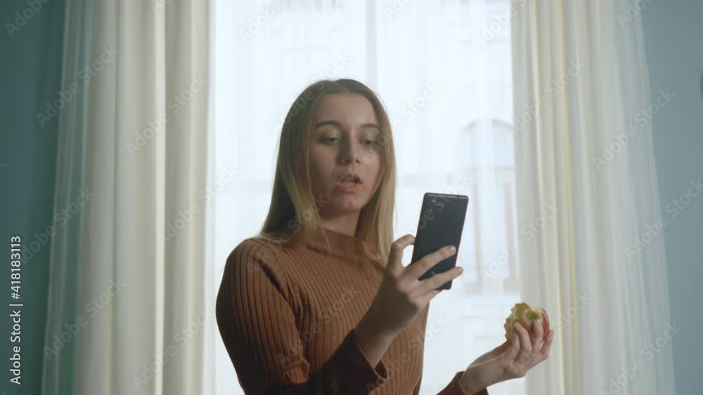 A young woman in blue jeans and a brown sweater sits on a beige sofa on the background of the window, biting a red apple and looking at something on the phone. The average plan. A young woman in blue jeans and a brown sweater sits on a beige sofa on the background of the window, biting a red apple and looking at something on the phone. The average plan.