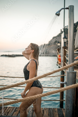 A beautiful young attractive woman in a black swimsuit with a beautiful figure stands under a summer shower on the pier by the sea. She is enjoying her vacation. Selective focus © Дмитрий Ткачук