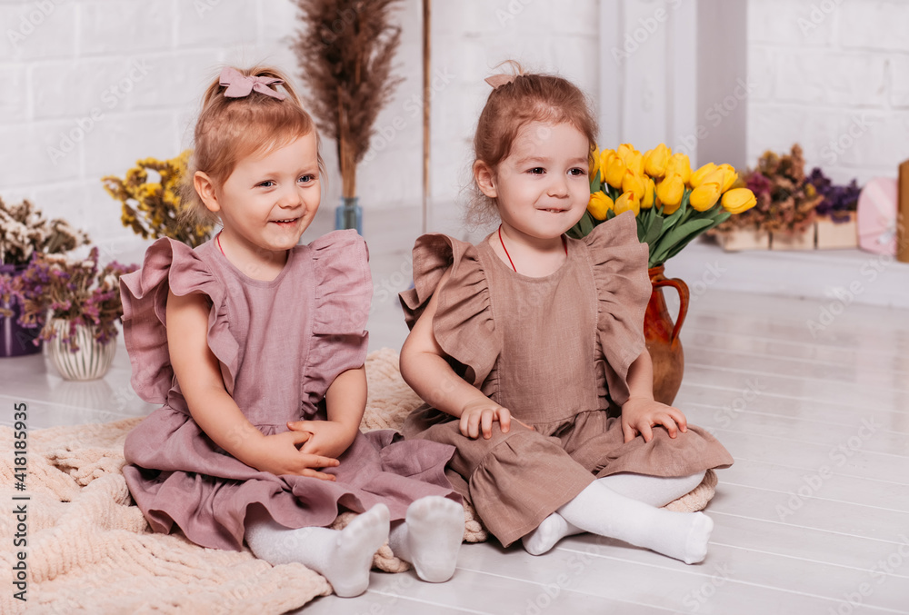Two little girls in identical dresses sit on the floor with flowers Two little girls in identical dresses sit on the floor with flowers