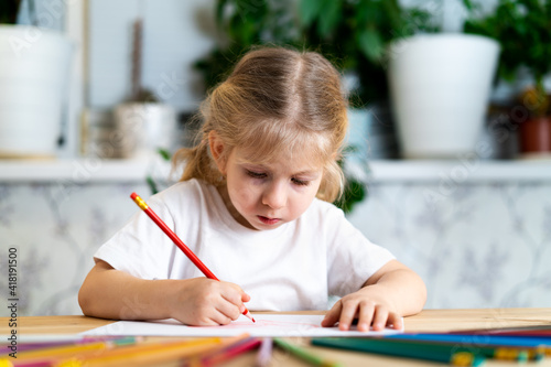 a little blonde girl is sitting at the table, smiling and drawing a heart with a red pencil, there are a lot of colored pencils on the table, learning concept