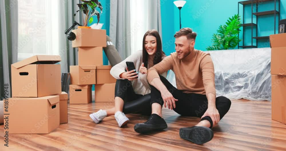 Young couple are using phone amid boxes in a new house. Teenage married couple at apartment web surfing on internet. Shopping online over cell phone. Online order. Shopping and payment online Young couple are using phone amid boxes in a new house. Teenage married couple at apartment web surfing on internet. Shopping online over cell phone. Online order. Shopping and payment online