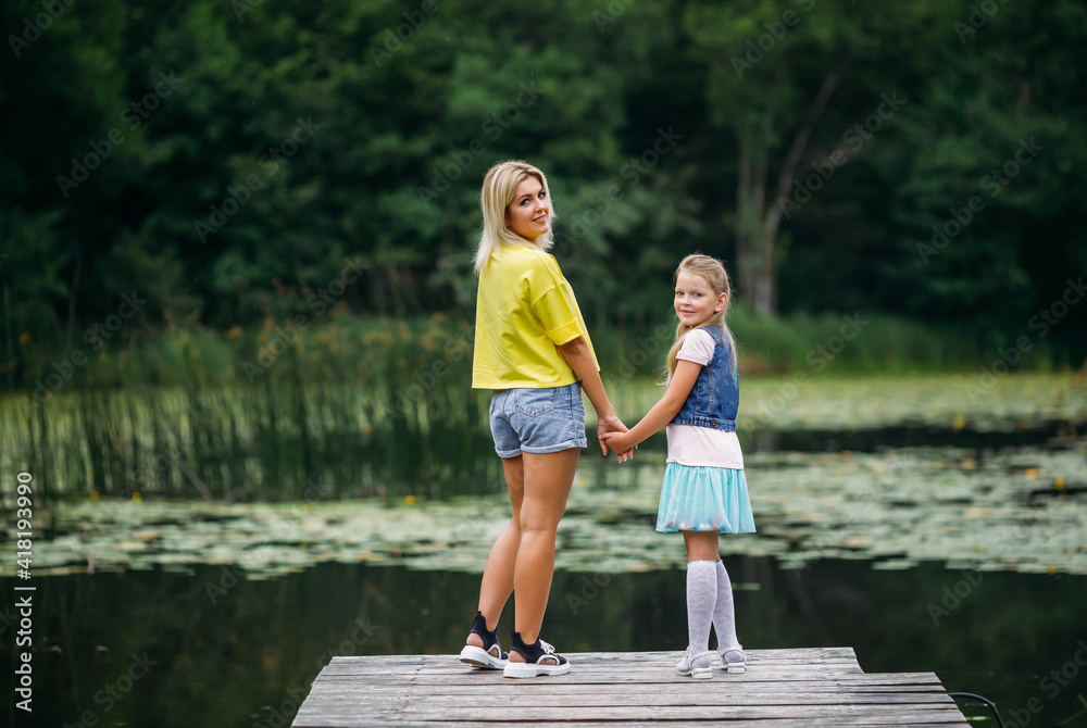 Young family, mother and daughter stand on a wooden pier holding hands, turning around and looking at the camera on a warm summer day Young family, mother and daughter stand on a wooden pier holding hands, turning around and looking at the camera on a warm summer day