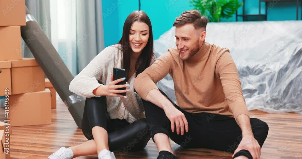 Happy young caucasian couple use digital smart phone discuss interior design renovation ideas sit on sofa on moving day, black new house renters owners tenants relax on couch with boxes having fun Happy young caucasian couple use digital smart phone discuss interior design renovation ideas sit on sofa on moving day, black new house renters owners tenants relax on couch with boxes having fun