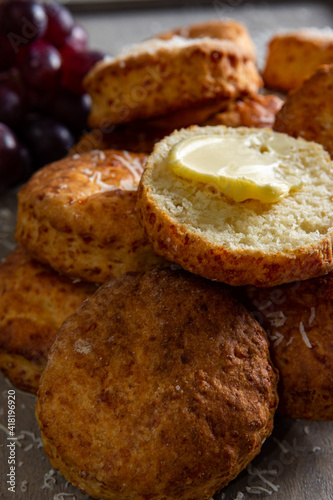 A close up view of a buttered cheese scone positioned atop others and dusted with parmesan cheese. Placed on a grey background with red grapes in the top left of the image.