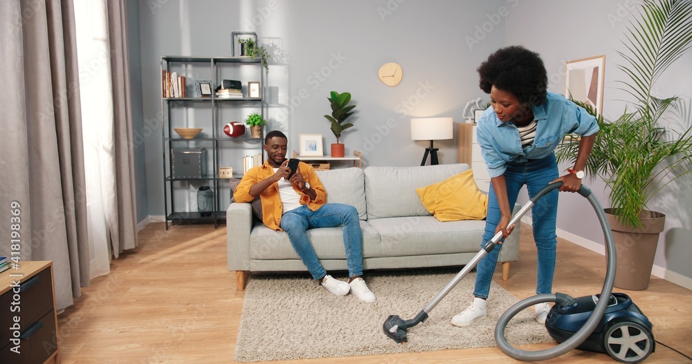 Happy beautiful young African American woman vacuuming carpet while handsome beloved man resting on couch texting on smartphone surfing online social network app, cleaning home concept