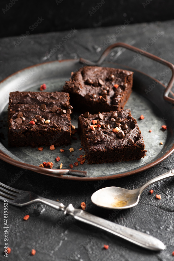 Dark cacao brownies with choco drops on old metal tray with vintage silverware on dark concrete background Dark cacao brownies with choco drops on old metal tray with vintage silverware on dark concrete background