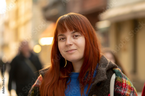 An attractive young woman with dyed red hair and a little overweight against the background of the city, a close-up street portrait. The cheerful face of a fat girl in daylight.