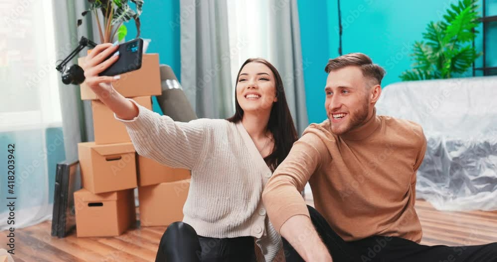 Happy couple taking selfie among cardboard boxes with things in new apartment. Handsome caucasian teenage girl holding smartphone. People grimace at camera. Happy couple taking selfie among cardboard boxes with things in new apartment. Handsome caucasian teenage girl holding smartphone. People grimace at camera.