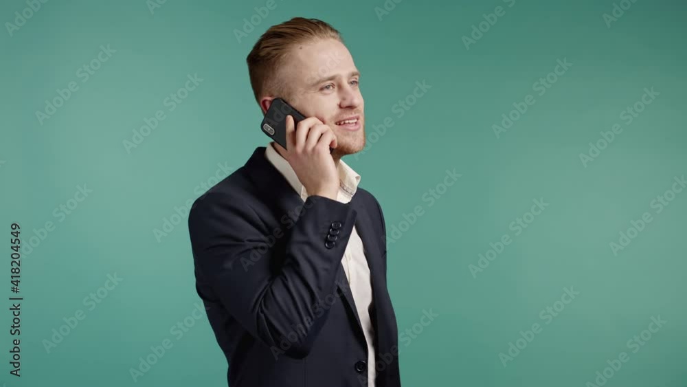 Young businessman speaks with smile on phone. Guy holding and using smartphone. Suit outfit. Blue studio background. Young businessman speaks with smile on phone. Guy holding and using smartphone. Suit outfit. Blue studio background.