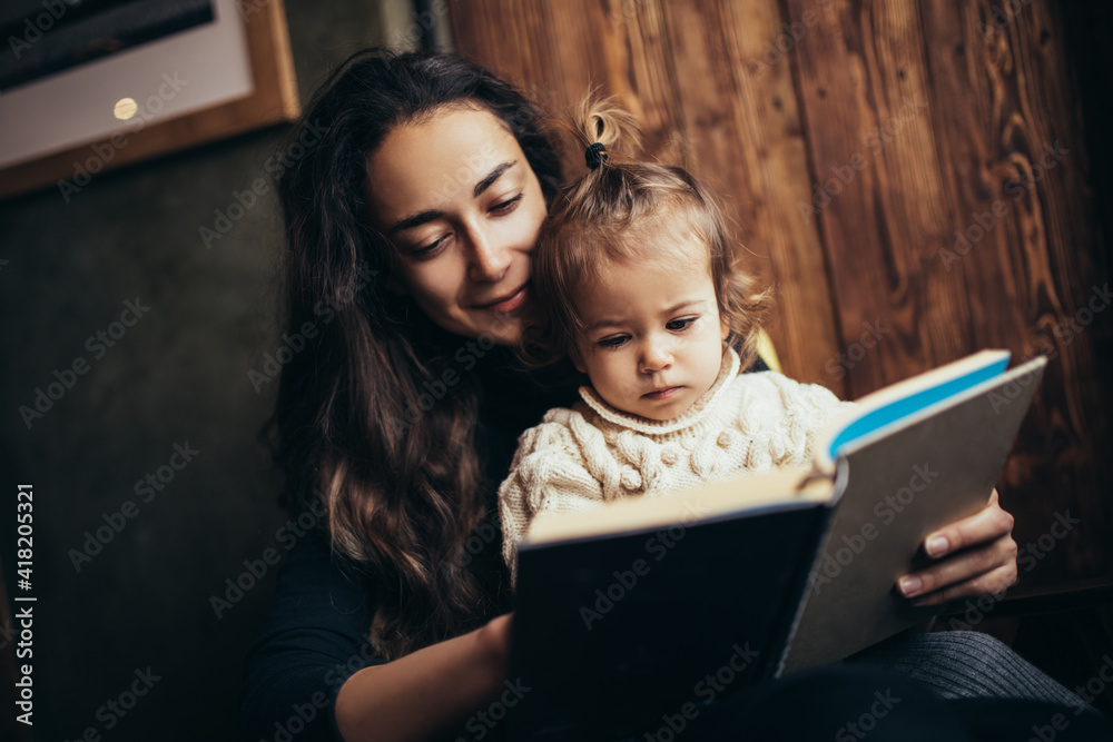 Young mother and her little daughter reading book together sitting in a comfortable chair.