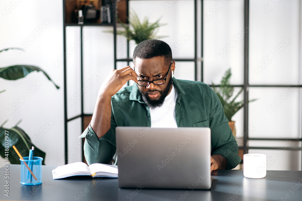 Focused dissatisfied multiracial black businessman, discouraged looks at the laptop, received unexpected message or news, sitting at workplace Focused dissatisfied multiracial black businessman, discouraged looks at the laptop, received unexpected message or news, sitting at workplace