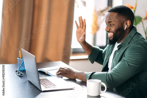 Online communication. Happy smiling African American guy, freelancer or student, waving hand, greeting, uses laptop for video conference with colleagues