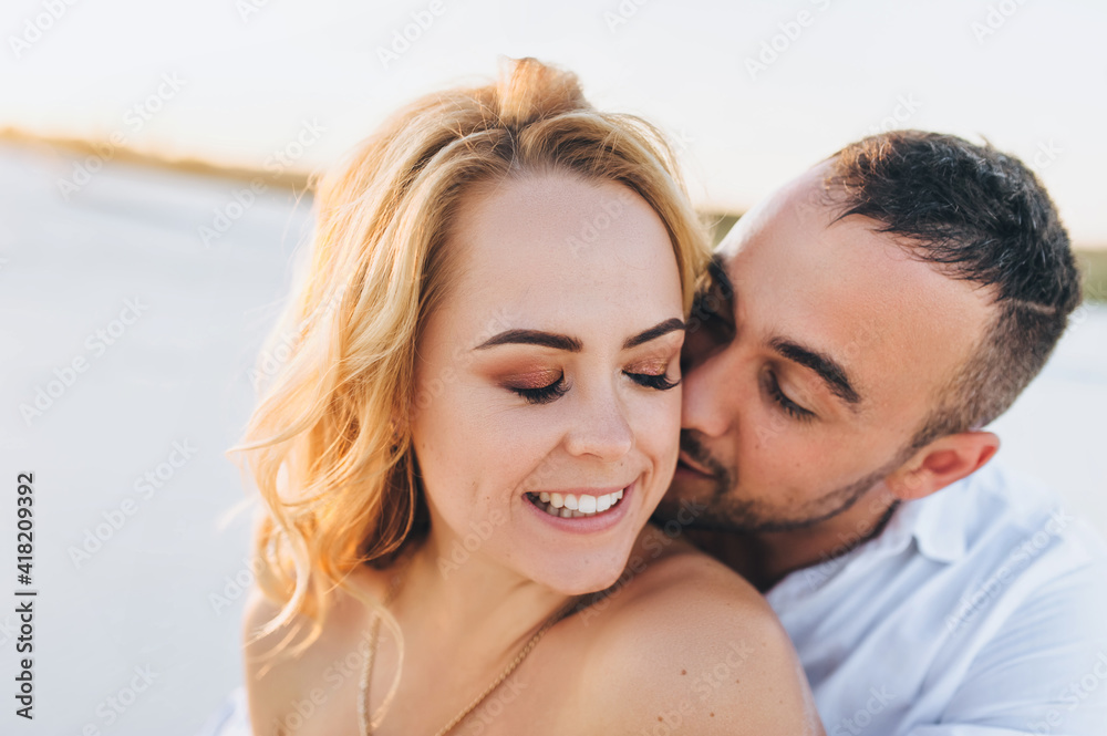 A young bearded man hugs a blonde against a background of white sand and gently kisses her neck. Portrait of emotional people at sunset. Love of newlyweds in the desert. Love story. A young bearded man hugs a blonde against a background of white sand and gently kisses her neck. Portrait of emotional people at sunset. Love of newlyweds in the desert. Love story.