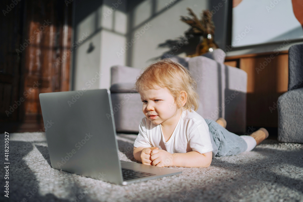 The child is lying on the floor resting and having a good time. A blonde girl is watching a children's program on a laptop, computer. Comfortable children's clothing. The child is lying on the floor resting and having a good time. A blonde girl is watching a children's program on a laptop, computer. Comfortable children's clothing.
