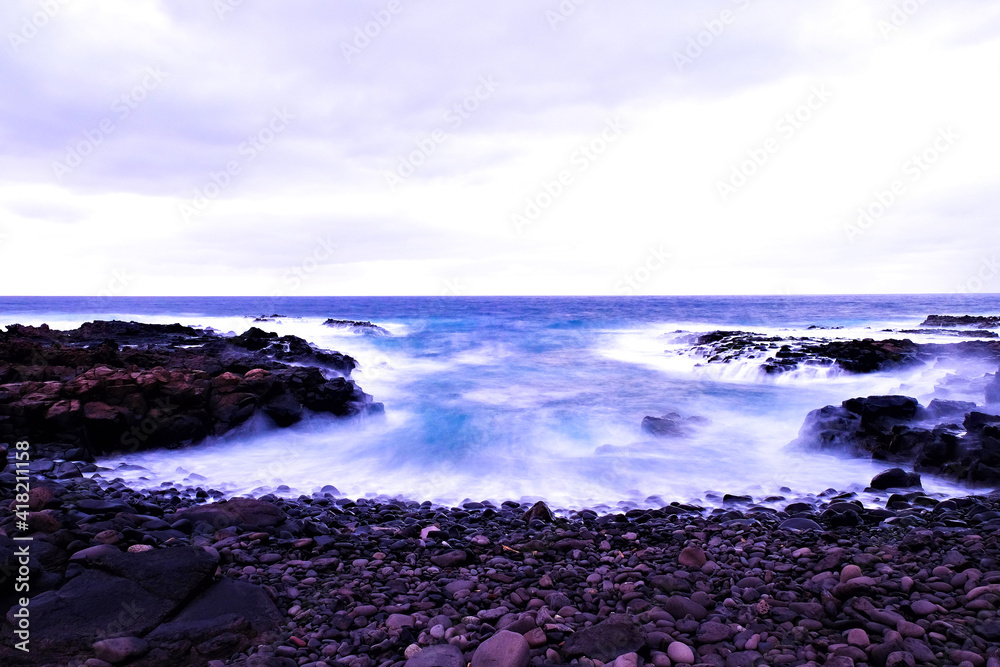 Waves filling a rocky islet after sunset, long exposure image with dreamy blue and white water in intentional blur.