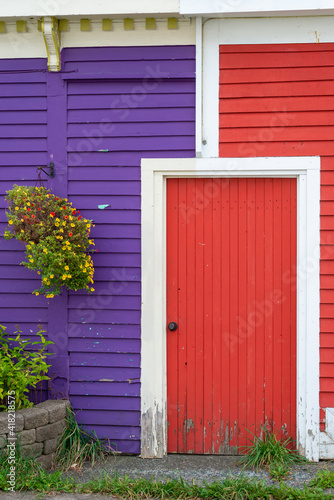A small red wooden common door with white wood trim around the exterior. The exterior door is in between a red wall and a purple wall. There's a hanging basket of flowers and a flower pot. 