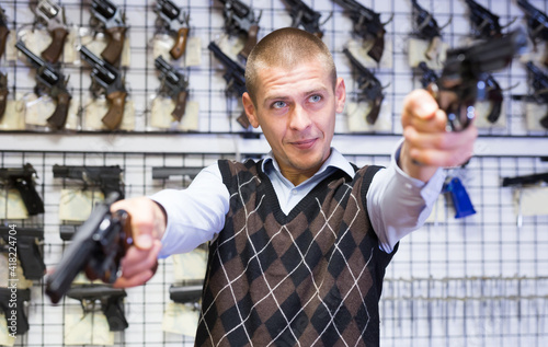 Portrait of focused adult man aiming with two pistols while choosing handguns in armory shop