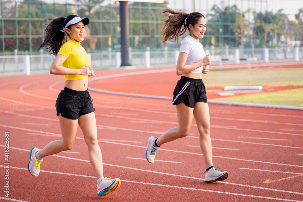 Two young Asian women in sports outfits jogging on running track in city stadium in the sunny morning to keep fitness and healthy lifestyle. Young fitness women run on the stadium track