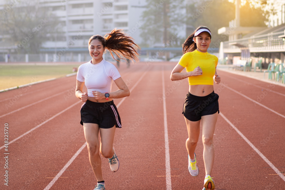 Obraz premium Two young Asian women in sports outfits jogging on running track in city stadium in the sunny morning to keep fitness and healthy lifestyle. Young fitness women run on the stadium track