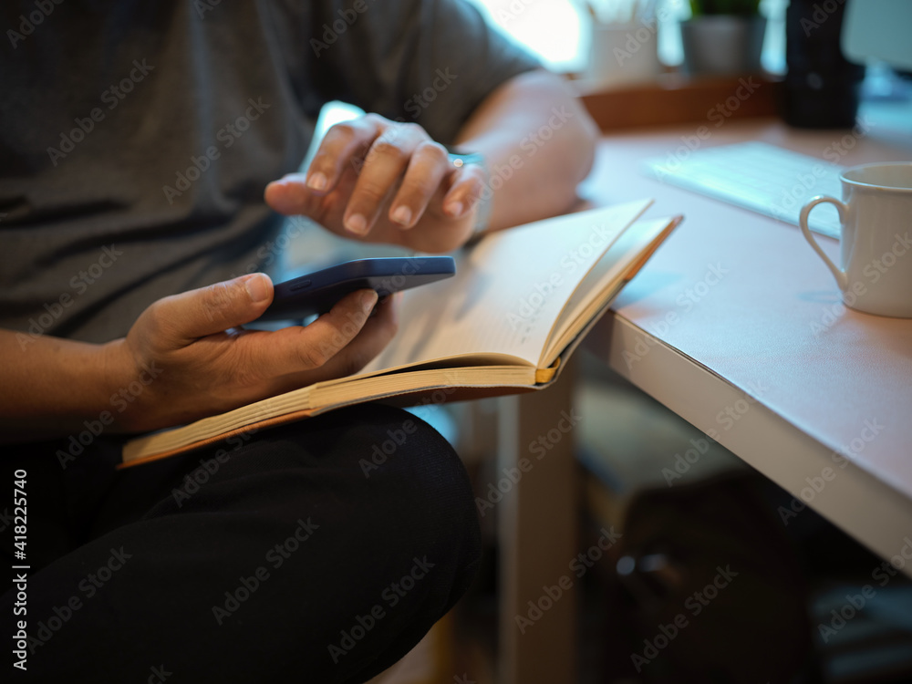 Male hand using smartphone while reading a book on computer desk in home office