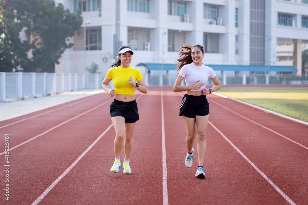 Obraz premium Two young Asian women in sports outfits jogging on running track in city stadium in the sunny morning to keep fitness and healthy lifestyle. Young fitness women run on the stadium track