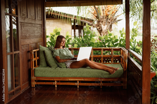 Young woman working on laptop on the couch at outdoor terrace at wooden house veranda with topical view