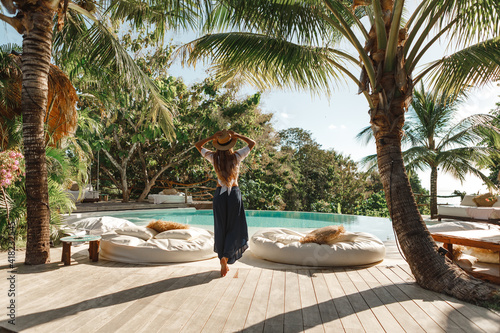 Young woman in straw hat walk near the swimming pool under palm tree. Tropical summer vacation concept