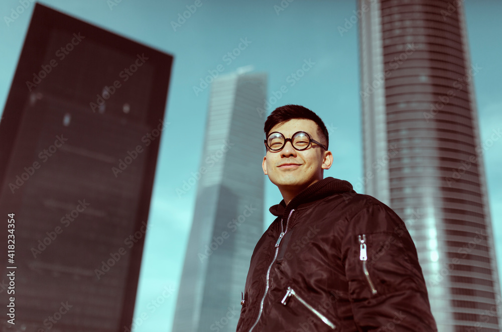 Chinese man, wearing round nerd glasses, with an friendly expression , with an skyscrapers in the background. Job, stock exchange, economy and chinese businessmen concept. Chinese man, wearing round nerd glasses, with an friendly expression , with an skyscrapers in the background. Job, stock exchange, economy and chinese businessmen concept.