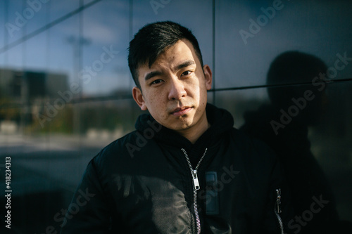 Portrait of a chinese man, wears urban clothing, looking at the camera with a serious expression. With an black wall of the street in the background. Job, unemployment and students concept. Copyspace