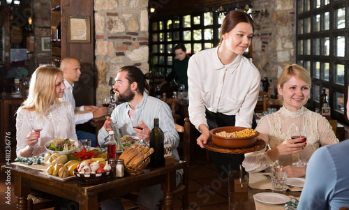 Polite waitress bringing ordered dishes to guests in comfortable country restaurant..