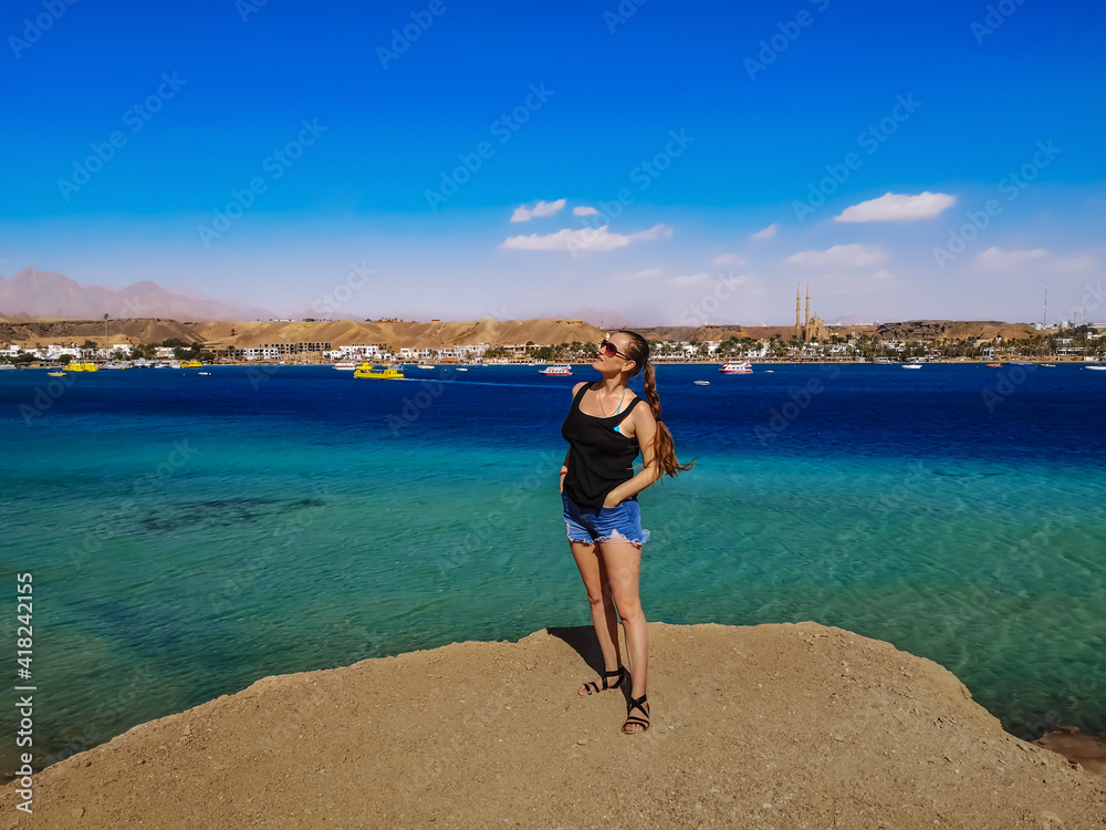 A young adult woman with braided hair stands on the edge of a cliff over the Red Sea in Sharm El Sheikh, Egypt. Cute slender european tourist posing against the backdrop of the sea panorama